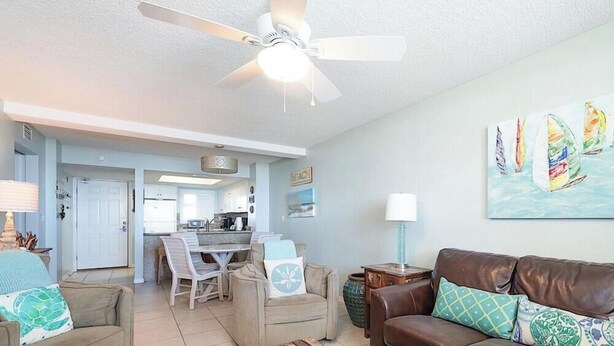 A spacious living room with a ceiling fan, a dining area with a kitchen in the background, and a painting of sailboats on the wall. - Sunswept 107 by Liquid Life (Orange Beach)