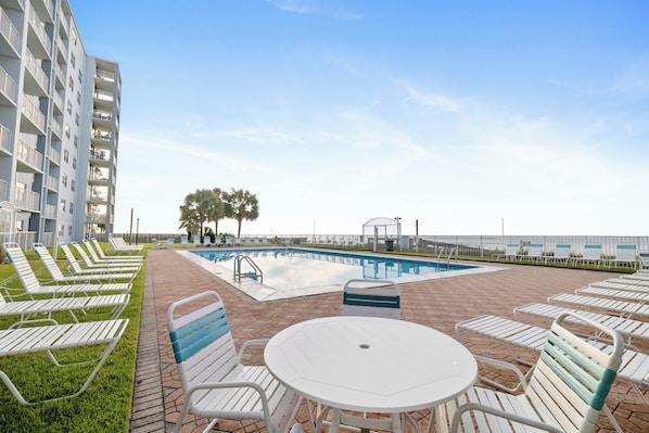 A pool area with lounge chairs and a table, surrounded by a building with balconies. - Sunswept 108 by Liquid Life (Orange Beach)