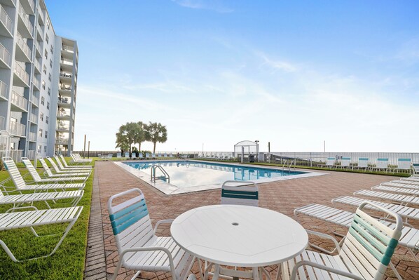 A pool area with lounge chairs and a table, surrounded by a building with balconies. - Sunswept 202 by Liquid Life (Orange Beach)