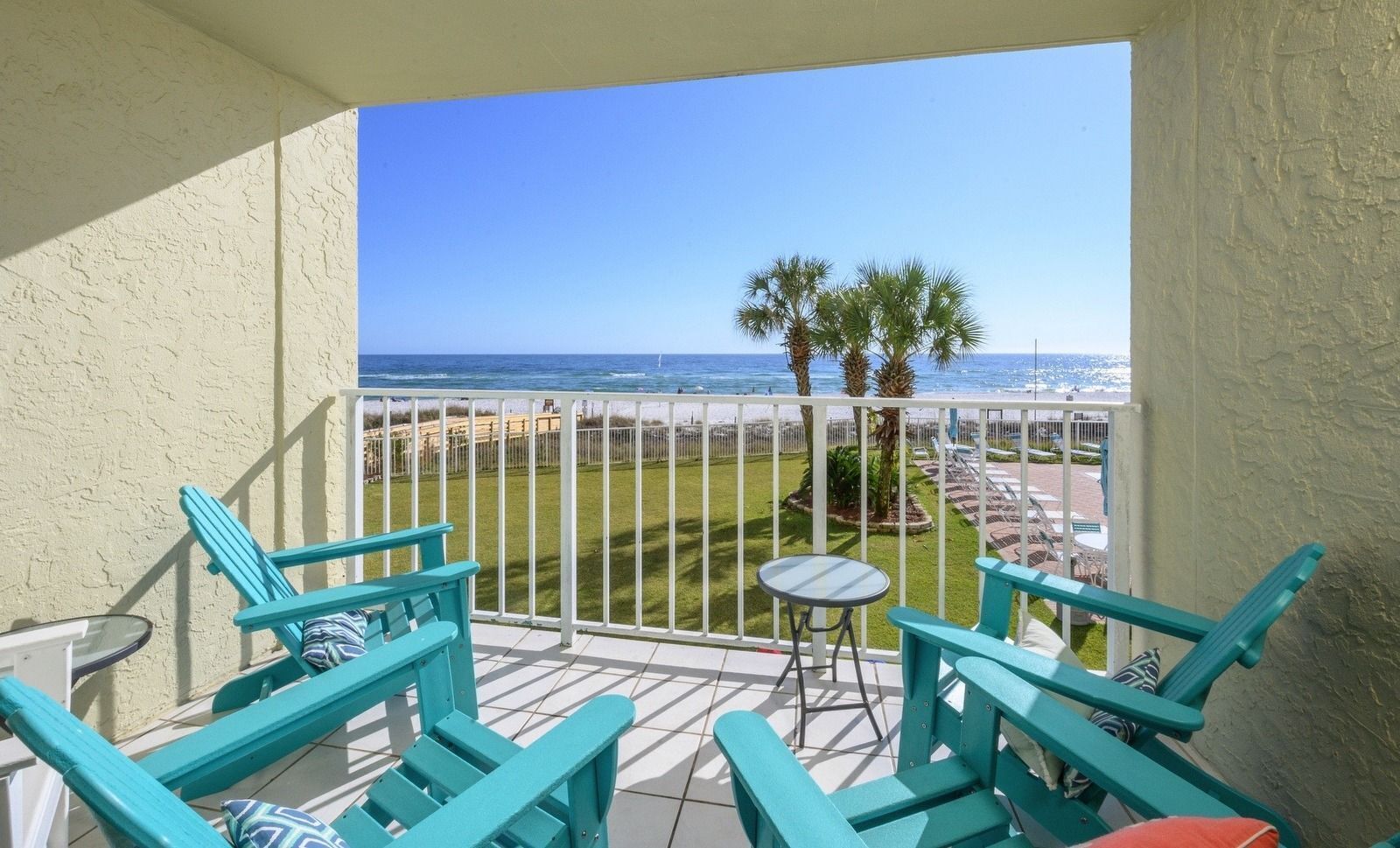 Ein Balkon mit türkisfarbenen Stühlen und einem kleinen Tisch mit Blick auf einen Strand mit Palmen und das Meer.