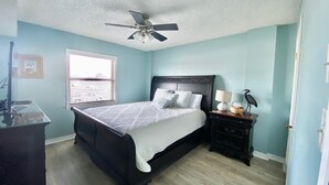 A bedroom with a bed, a nightstand, a ceiling fan, a window with blinds, and a door. - Sunswept 202 by Liquid Life (Orange Beach)