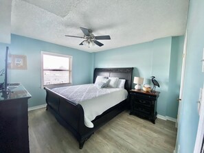 A bedroom with a bed, a nightstand, a ceiling fan, a window with blinds, and a door. - Sunswept 202 by Liquid Life (Orange Beach)