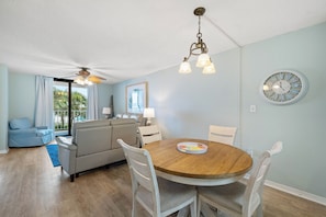 A living room with a round wooden table, white chairs, a sofa, and a ceiling fan. - Sunswept 202 by Liquid Life (Orange Beach)