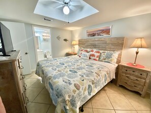 Une chambre à coucher comprenant un lit, une table de chevet, une lampe, un ventilateur de plafond, un tableau et une porte donnant sur une salle de bain.