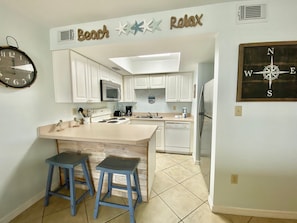 A kitchen with white cabinets, a microwave, stove, and a refrigerator. There are two blue stools and a clock on the wall. - Sunswept 204 by Liquid Life (Orange Beach)