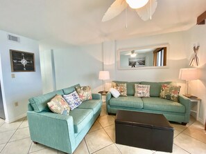 A living room with two teal sofas, a black ottoman, and a wall-mounted lamp. - Sunswept 204 by Liquid Life (Orange Beach)