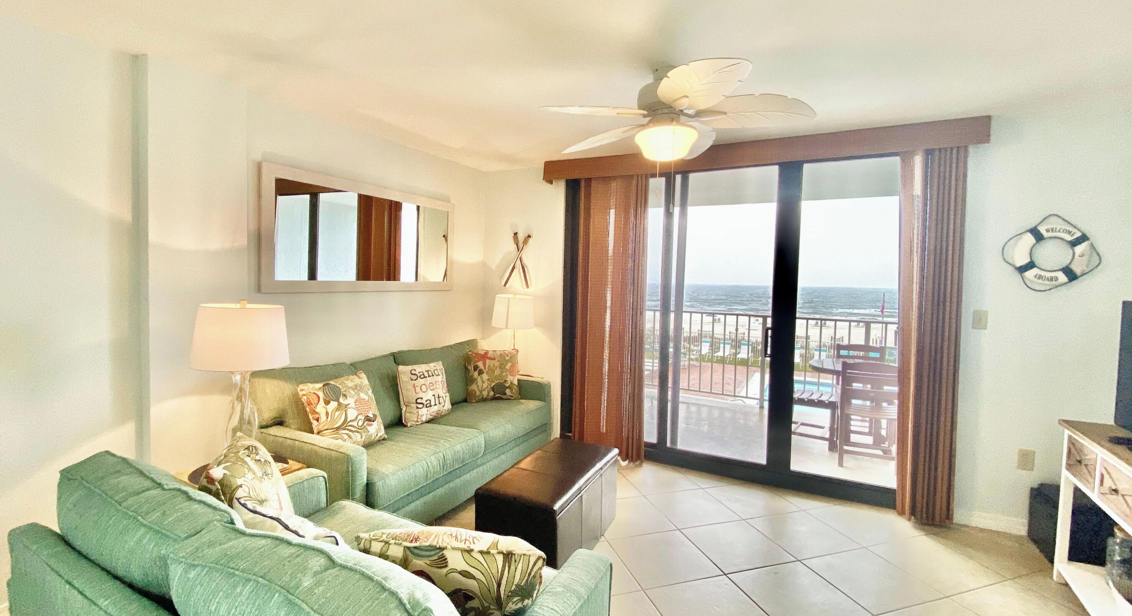 A living room with a green sofa, a ceiling fan, a view of the ocean through a sliding glass door, and a 'Welcome Aboard' sign.
