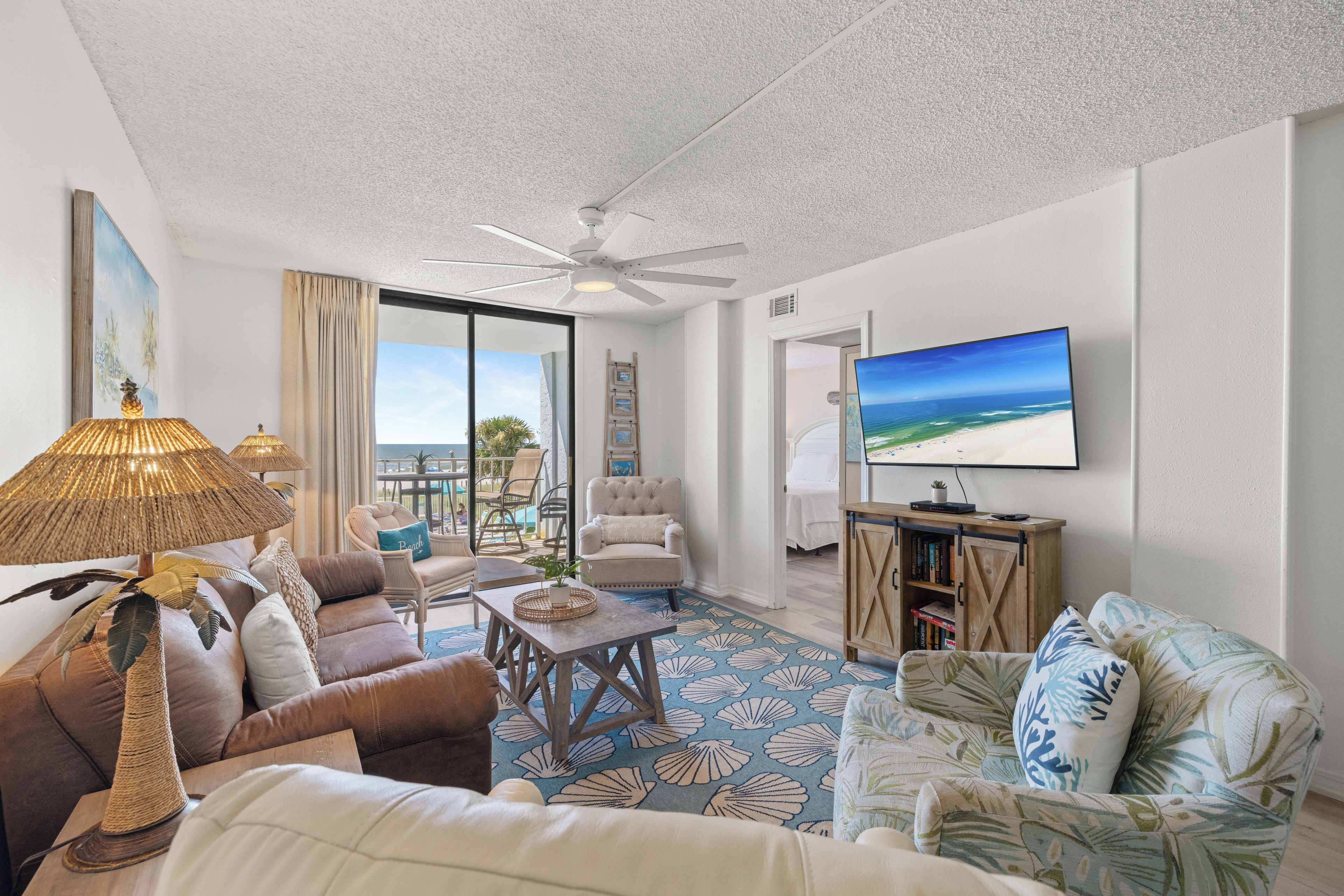A spacious living room with a ceiling fan, a television mounted on the wall, and a view of the ocean through large windows.