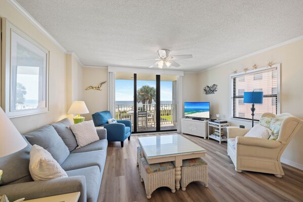 Ein Wohnzimmer mit einer Couch, Sesseln, einem Couchtisch, einem Fernseher und Blick auf den Strand durch große Fenster.