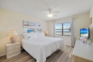 Habitación de hotel con cama, escritorio, ventilador de techo, ventana con vista a la playa y un cuadro en la pared.