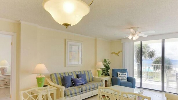 A bright living room with a sofa, two armchairs, a coffee table, and a ceiling fan. There is a large window providing a view of the outdoors. - Sunswept 208 by Liquid Life (Orange Beach)