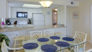 A modern kitchen with white cabinets, a microwave, and a refrigerator. There is a glass dining table with blue cushions and white wicker chairs. - Sunswept 208 by Liquid Life (Orange Beach)