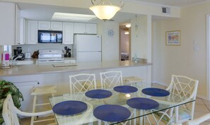 A modern kitchen with white cabinets, a microwave, and a refrigerator. There is a glass dining table with blue cushions and white wicker chairs. - Sunswept 208 by Liquid Life (Orange Beach)