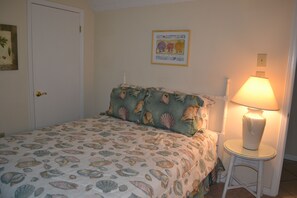 A bedroom with a bed covered in a seashell patterned bedspread, a bedside lamp, and a framed picture on the wall. - Sunswept 208 by Liquid Life (Orange Beach)