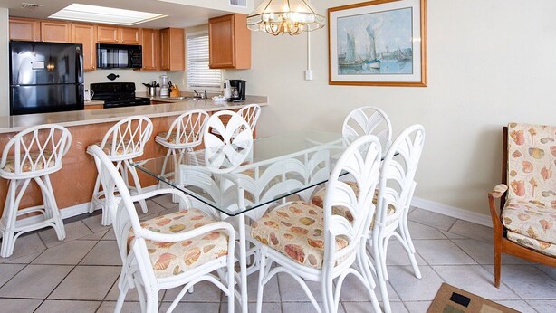 Cocina equipada con refrigerador, microondas, estufa y lavadero. Hay una zona de comedor con una mesa de vidrio y sillas blancas. Un cuadro adorna la pared.