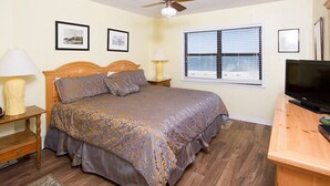 Ein Schlafzimmer mit einem Bett, zwei Nachttischlampen, einem Fernseher, einem Fenster mit Jalousien und zwei gerahmten Bildern an der Wand.