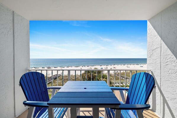 Ein Balkon mit Tisch und zwei Stühlen, der Blick auf einen Strand mit Sonnenschirmen und Menschen fällt.