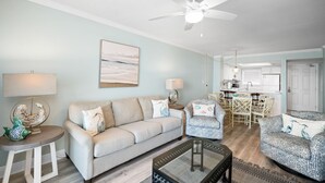 A living room with a sofa, two armchairs, a coffee table, a ceiling fan, and a kitchen area in the background. - Sunswept 402 by Liquid Life (Orange Beach)