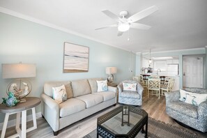 A living room with a sofa, two armchairs, a coffee table, a ceiling fan, and a kitchen area in the background. - Sunswept 402 by Liquid Life (Orange Beach)