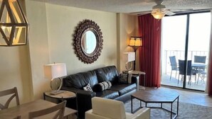 A hotel room with a black leather sofa, a round mirror, a lamp, a coffee table, and a view of the outside. - Sunswept 403 by Liquid Life (Orange Beach)