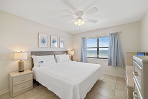 Une chambre d’hôtel avec un lit, des tables de chevet, un ventilateur de plafond, des œuvres d’art au mur, une fenêtre avec des rideaux et une vue sur la plage.