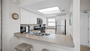A modern kitchen with white cabinets, a stainless steel refrigerator, a microwave, and a coffee maker. - Sunswept 406 by Liquid Life (Orange Beach)