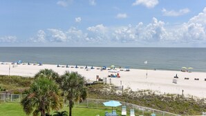 A beach with umbrellas and lounge chairs, a fenced area with palm trees, and a clear view of the ocean. - Sunswept 406 by Liquid Life (Orange Beach)