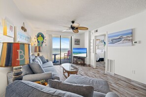 Un salon avec un canapé, un téléviseur et une vue sur la plage, visible à travers les portes vitrées.