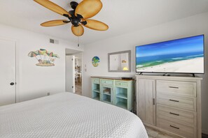 Une chambre à coucher avec un lit, un ventilateur de plafond, un téléviseur diffusant une scène de plage et une porte donnant sur une autre pièce.