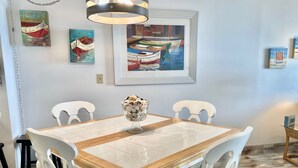 Un coin repas avec une table en bois et des chaises blanches, un vase contenant des fleurs séchées et des tableaux accrochés au mur.