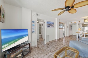 A spacious living area with a television displaying a beach scene, a ceiling fan, and a dining area with a table and chairs. - Sunswept 407 by Liquid Life (Orange Beach)