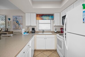 A modern kitchen with white cabinetry, a large island with a sink, and a window with blinds. - Sunswept 408 by Liquid Life (Orange Beach)