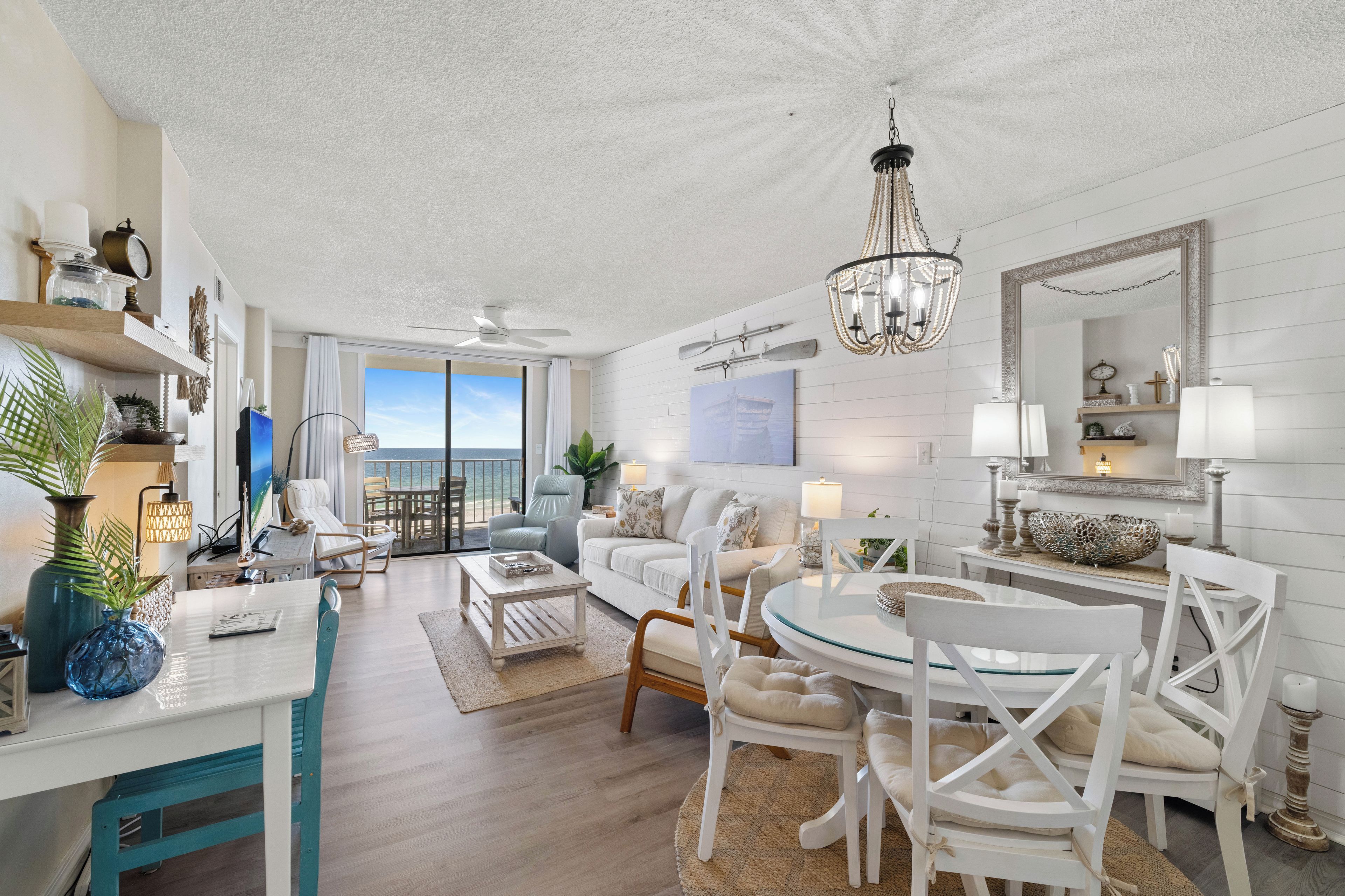 Amplia sala de estar con mesa de comedor, mobiliario blanco, lámpara de araña y un ventanal con vista al océano.