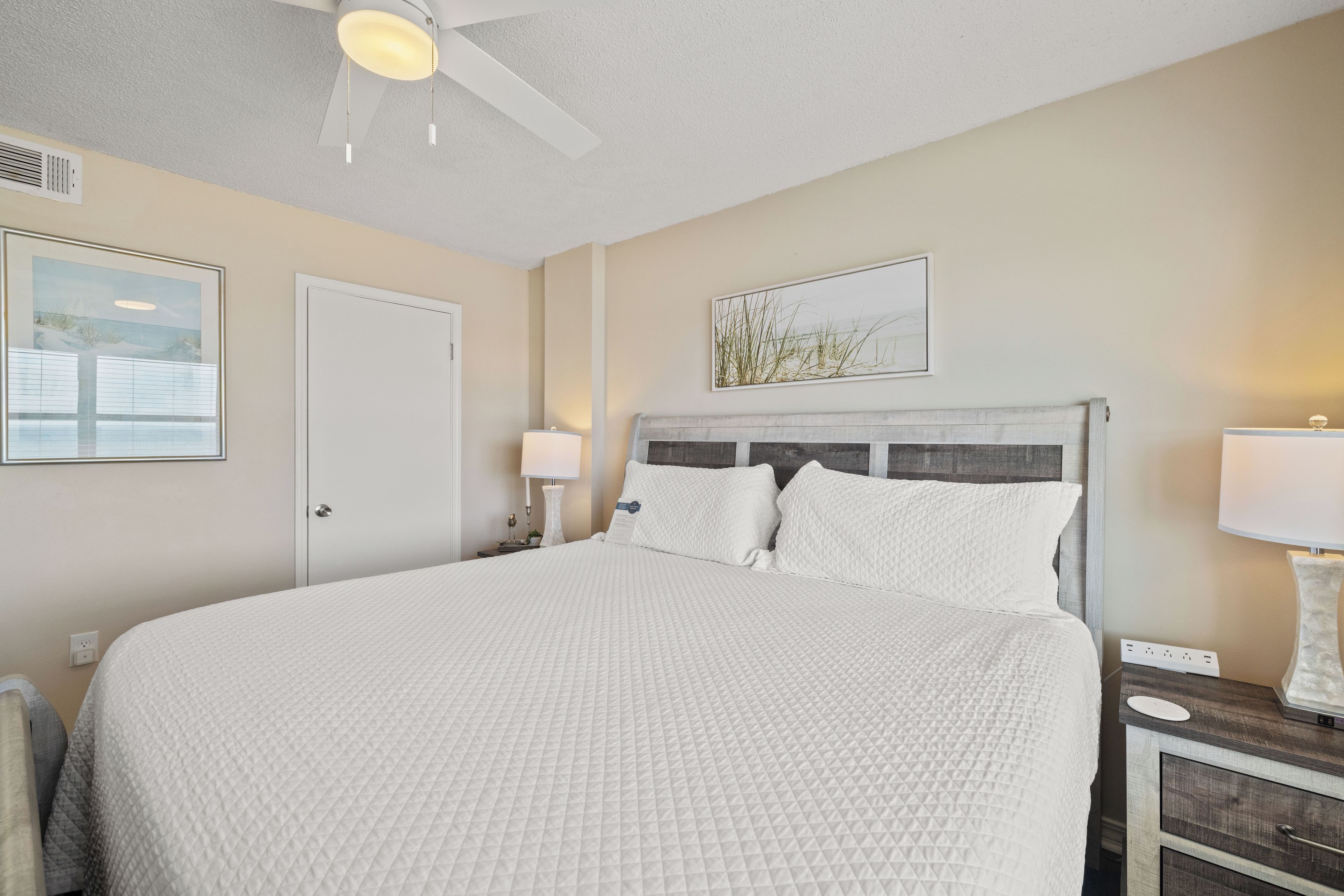 Une chambre d’hôtel avec un grand lit, un ventilateur de plafond, une table de chevet avec une lampe et des œuvres d’art accrochées aux murs.