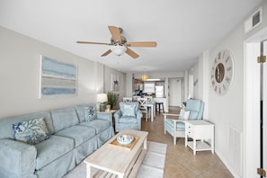 Ein Wohnzimmer mit Sofa, Couchtisch und Deckenventilator. Im Hintergrund ist eine Küche mit Essbereich zu sehen.