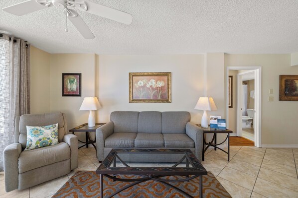 A living room with a grey sofa, a coffee table, a chair, two lamps, and a ceiling fan. - Sunswept 601 by Liquid Life (Orange Beach)