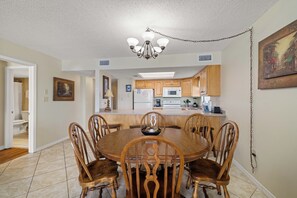 A dining area with a wooden table and chairs, a chandelier, and a kitchen in the background. - Sunswept 601 by Liquid Life (Orange Beach)