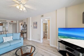 A living room with a blue sofa, a glass coffee table, a ceiling fan, a TV displaying a beach scene, and a kitchen in the background. - Sunswept 602 by Liquid Life (Orange Beach)