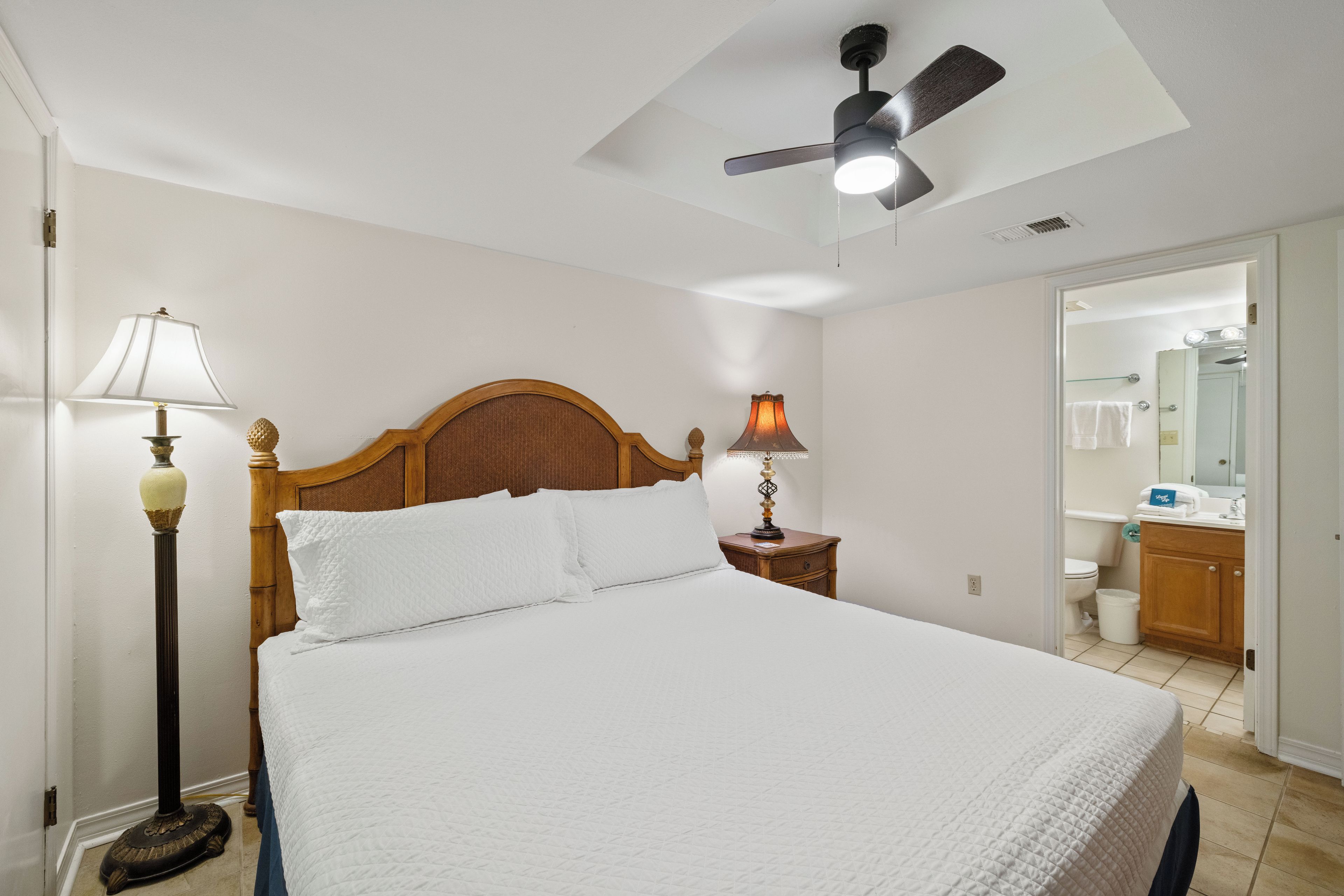 Une chambre  à coucher avec un lit, deux lampes de chevet, un ventilateur de plafond et une salle de bain visible à travers une porte ouverte.
