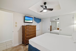 Ein Schlafzimmer mit Bett, Kommode, Deckenventilator, einem an der Wand befestigten Fernseher und einem Fenster mit Jalousien.