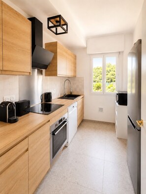 A modern kitchen with wooden cabinets, stainless steel appliances, and a window with a view of greenery. - Val de Cagnes Residence - Stylish and Peaceful – Sleeps 6 – Air Conditioning (Cagnes-sur-Mer)