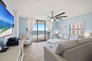 A living room with a large TV, a ceiling fan, a sofa, a coffee table, and a balcony with a view of the beach. - Sunswept 608 by Liquid Life (Orange Beach)