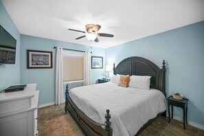 Ein Schlafzimmer mit Bett, Schreibtisch, Nachttisch, Deckenventilator und einem Fenster mit Jalousien.