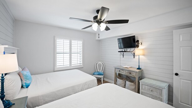 Ein Schlafzimmer mit zwei Betten, einem Deckenventilator, einem Fernseher und einem Schreibtisch mit Lampe.