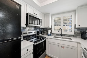 A modern kitchen with white cabinetry, a black refrigerator, stainless steel appliances, and a window with blinds. - Sunswept 701 by Liquid Life (Orange Beach)