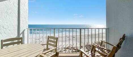 A balcony with a view of the beach and ocean. There is a wooden table and chairs.