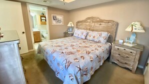 A bedroom with a bed, two nightstands, a ceiling fan, a door leading to a bathroom, and a small table with a lamp. - Sunswept 704 by Liquid Life (Orange Beach)