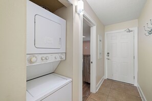 A laundry room with a front-loading washer and dryer, a door leading to another room, and a wall-mounted light fixture. - Sunswept 704 by Liquid Life (Orange Beach)