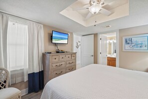 Une chambre à coucher comprenant un lit, une commode, un téléviseur, un ventilateur de plafond et une porte donnant sur une salle de bain.