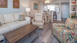 Een woonkamer met een bank, een salontafel, een stoel en een fauteuil met een patroon. Achtergrond toont een eethoek met een tafel en stoelen.